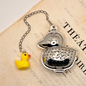 Pode incluir: Um infusor de chá de metal prateado em forma de pato com um amuleto de pato de borracha amarelo preso à corrente. O infusor tem um fundo de malha e é projetado para conter folhas de chá soltas.