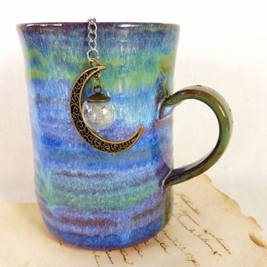 Könnte beinhalten: Eine blaue und grüne Keramiktasse mit einem Halbmond-Anhänger, der am Henkel hängt. Der Anhänger ist aus Metall und hat eine kleine Glaskugel daran.