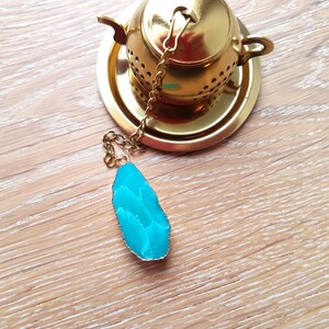Peut inclure: Une infuseur &agrave; th&eacute; en or avec une pierre d'agate turquoise attach&eacute;e &agrave; la cha&icirc;ne. L'infuseur est pos&eacute; sur une soucoupe dor&eacute;e.