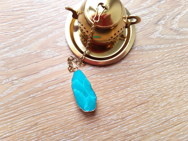 Peut inclure: Une infuseur &agrave; th&eacute; en or avec une pierre d'agate turquoise attach&eacute;e &agrave; la cha&icirc;ne. L'infuseur est pos&eacute; sur une soucoupe dor&eacute;e.
