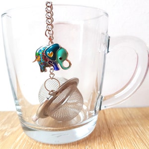 May include: A colourful elephant tea infuser with a heart on its trunk, hanging from a chain inside a clear glass mug.