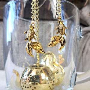 Puede incluir: Infusor de té dorado con dos colgantes en forma de pez dorado que cuelgan de cadenas. El infusor está en una taza de vidrio transparente.
