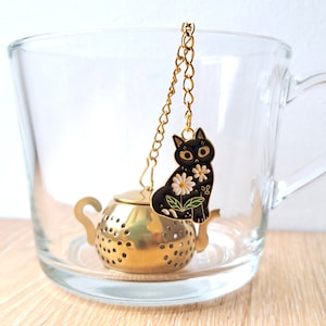 Könnte beinhalten: Goldener Teesieb in Teekannenform mit einem schwarzen Katzenanhänger mit weißen Blumen. Der Teesieb hängt an einer goldenen Kette in einer durchsichtigen Glas Tasse.