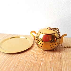 Peut inclure: Infuseur &agrave; th&eacute; en forme de th&eacute;i&egrave;re en m&eacute;tal dor&eacute; avec un couvercle et une cha&icirc;ne. L'infuseur est pos&eacute; sur une plaque en m&eacute;tal dor&eacute;.