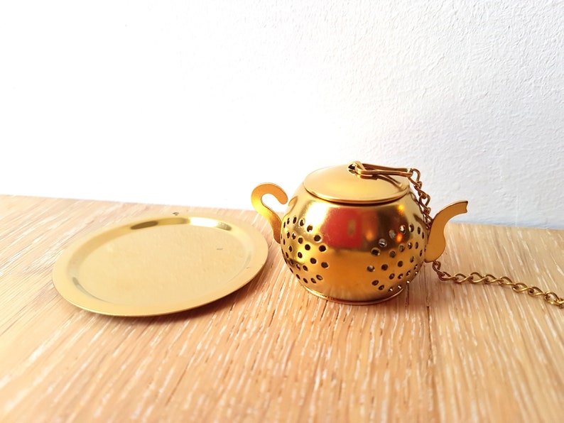 Peut inclure: Infuseur &agrave; th&eacute; en forme de th&eacute;i&egrave;re en m&eacute;tal dor&eacute; avec un couvercle et une cha&icirc;ne. L'infuseur est pos&eacute; sur une plaque en m&eacute;tal dor&eacute;.