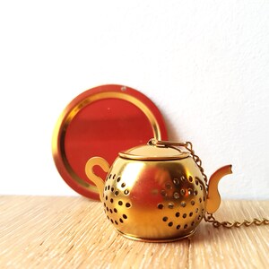 Peut inclure: Infuseur &agrave; th&eacute; en m&eacute;tal dor&eacute; en forme de th&eacute;i&egrave;re avec une cha&icirc;ne attach&eacute;e. L'infuseur a un corps rond avec de petits trous et un bec verseur. L'infuseur est pos&eacute; sur une surface en bois.