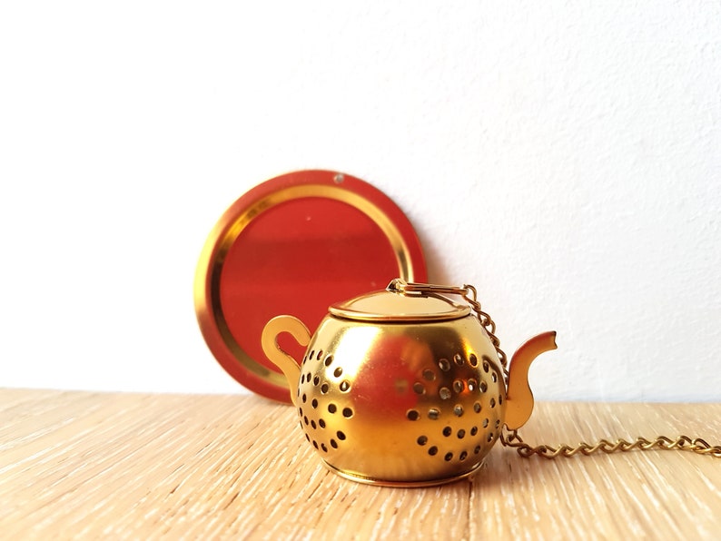 Peut inclure: Infuseur &agrave; th&eacute; en m&eacute;tal dor&eacute; en forme de th&eacute;i&egrave;re avec une cha&icirc;ne attach&eacute;e. L'infuseur a un corps rond avec de petits trous et un bec verseur. L'infuseur est pos&eacute; sur une surface en bois.
