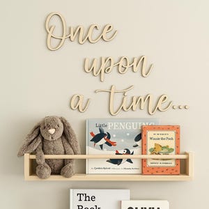 May include: Wooden shelves with children's books and a stuffed bunny. The top shelf holds "Little Penguin" and "Winnie the Pooh" books. The bottom shelf displays "The Book With No Pictures" and "Olivia". The words "Once upon a time..." are above the shelves.