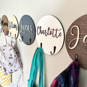 Peut inclure: Une série de plaques murales circulaires en bois avec des prénoms découpés au laser dans diverses polices et couleurs. Chaque plaque est équipée de deux crochets en métal noir pour suspendre des objets. Les prénoms sont Poppy, Mason, Oliver, Lucas, Charlotte et Jack.