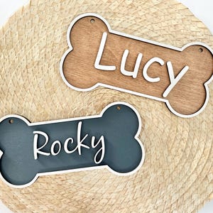 May include: Two bone-shaped wooden signs with white borders and small holes for hanging. One sign is natural light brown wood with the white text Lucy, and the other is dark blue-grey with the white text Rocky in a script font.