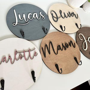 May include: Five circular wooden wall plaques, each featuring a unique name in raised script lettering and two black metal hooks. The plaques come in various wood finishes including dark gray, light natural, medium brown, and white with pink text.