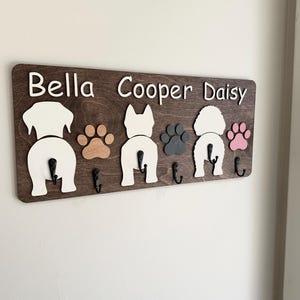 May include: Wooden key holder with the names "Bella", "Cooper", and "Daisy" in white lettering. The holder features three dog silhouettes and paw prints in various colors. Each dog has a black hook below.
