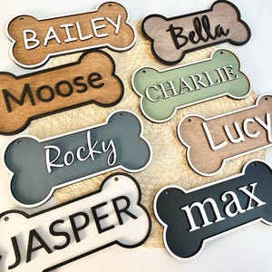 May include: A collection of wooden dog bone-shaped name tags. Each tag features a different name in a contrasting color, including "Bailey," "Bella," "Moose," "Charlie," "Rocky," "Lucy," "Jasper," and "Max." The tags have a layered design with a contrasting border.
