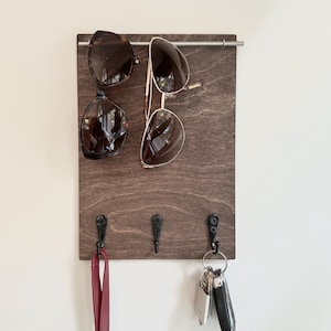 May include: A wooden sunglasses and key holder with a dark brown finish. Two pairs of sunglasses hang from a metal bar, and a red leather leash and keys hang from the hooks below. The holder is mounted on a white wall.