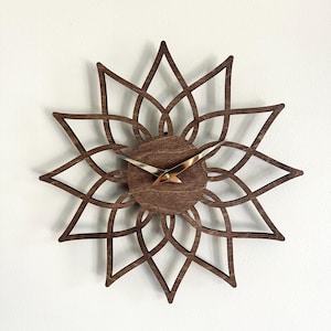Peut inclure: Une horloge murale en bois avec un motif en forme d'éclat d'étoile. L'horloge présente une finition marron foncé, un centre circulaire et des aiguilles dorées. Le design de l'horloge est complexe et géométrique.