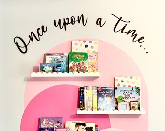 May include: Three white scalloped floating shelves mounted on a wall with a pink painted arch design. The shelves hold various children's books with colorful covers. Above the shelves, the black cursive text reads Once upon a time...