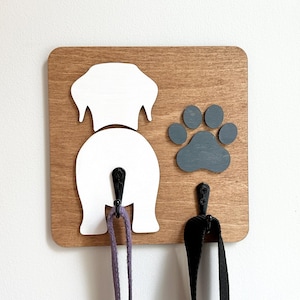 May include: A square wooden wall-mounted hook rack featuring a white silhouette of a dog's rear end and a dark grey paw print. Two black metal hooks are attached to the wood, with one hook positioned on the dog's tail and one below the paw.