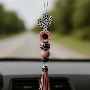 May include: A car charm featuring a black and white spotted cactus, a pink flower, zebra-striped bead, pink and black beads, and a pink tassel. The charm hangs from a white string, adding a decorative touch to any vehicle.