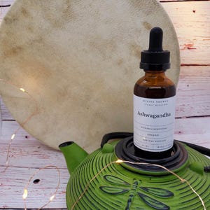 May include: A brown glass bottle of Ashwagandha herbal supplement with a black dropper, resting on a green teapot with a dragonfly design. A large, round, tan drum is in the background. The label reads "Divine Source Plant Healing".