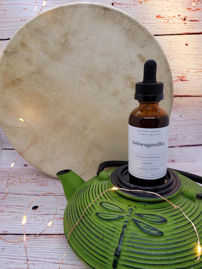 May include: A brown glass bottle of Ashwagandha herbal supplement with a black dropper, resting on a green teapot with a dragonfly design. A large, round, tan drum is in the background. The label reads "Divine Source Plant Healing".