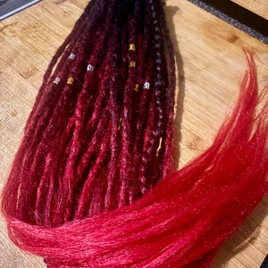 May include: A bundle of red and black ombre dreadlocks with silver and gold hair beads. The dreadlocks are arranged on a wooden chopping board. The hair is a deep red colour.