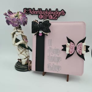 Op de afbeelding: Een roze planner met zwarte en glitterende strikken, een zwarte band en de tekst "100 Mystery Envelope Challenge". Links staat een decoratief skeletfiguur met een paarse bloem. De planner staat op een kleine houten standaard.