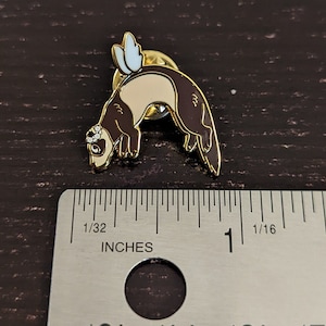 May include: A gold-colored metal pin featuring a ferret design. The ferret is brown and tan with a small floral crown and light blue wings. The pin measures approximately 1 inch in length.