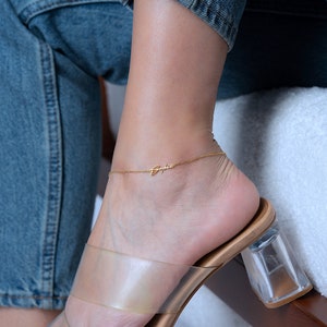 Può includere: Un braccialetto dorato per la caviglia con il nome "Sophia" inciso su di esso. Il braccialetto è indossato sulla caviglia di una persona con un sandalo a cinturino trasparente.