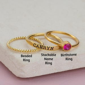 Puede incluir: Tres anillos de oro apilados uno encima del otro. El anillo inferior es un anillo de cuentas. El anillo del medio es una banda delgada con el nombre "CAMRYN" grabado en él. El anillo superior es una banda delgada con una piedra preciosa rosa.
