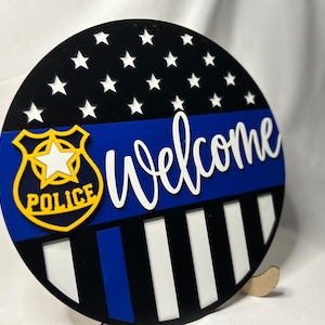 May include: Round wooden sign with a black background and white stars. Features a blue stripe, a police badge, and the word "Welcome" in white script. The bottom has black and white stripes.