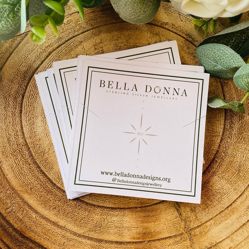 K&ouml;nnte beinhalten: Ein Stapel wei&szlig;er Schmuck-Displaykarten mit schwarzen R&auml;ndern. Die Karten zeigen den Text "Bella Donna Sterling Silver Jewellery" und einen stilisierten Sternenausbruch mit den Buchstaben "BD" in der Mitte. Die Unterseite der Karte enth&auml;lt die Website-Adresse "www.belladonnadesigns.org" und den Social-Media-Handle "@belladonnadesignjewellery".