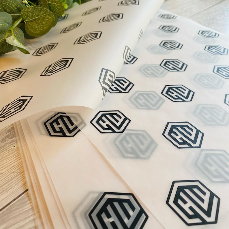 May include: Translucent white wrapping paper with a repeating black geometric logo pattern. The paper is partially rolled, revealing multiple layers. A few green leaves are visible in the upper left corner.