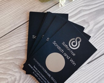 May include: A stack of black scratch-off cards with a silver logo and the text "Saffron hair Scratch and Win". The cards have a silver circle in the center that can be scratched off to reveal a prize. The text "WWW.SAFFRONHAIRLTD.COM" and "Terms and Conditions Apply" are printed on the cards. The text "please email saffronhairltd@gmail.com to claim your prize" is also printed on the cards.