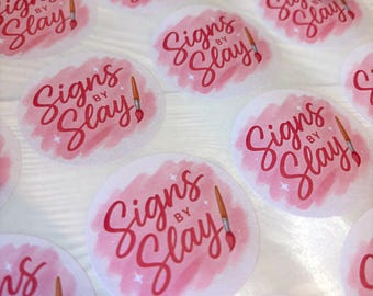 May include: A collection of round stickers with the text "Signs by Slay!" in a cursive font. The text is in red, set against a pink watercolor-style background. A paintbrush graphic is also included.