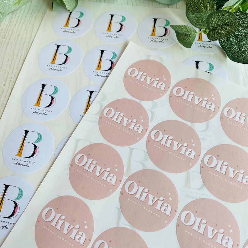 Op de afbeelding: Twee vellen met ronde stickers. E&eacute;n vel heeft witte stickers met een zwarte en groene letter B en de tekst "Ben Dantzon Photographer". Het andere vel heeft roze stickers met de tekst "Olivia Beauty Blogger" in wit.