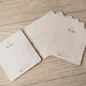 K&ouml;nnte beinhalten: Ein Stapel wei&szlig;er Visitenkarten mit goldenen Flocken. Die Karten tragen den Schriftzug "Koko" in Schwarz. Der Text "925 Silver" und "www.kokojewellery.com" ist ebenfalls auf den Karten gedruckt, sowie Social-Media-Icons.