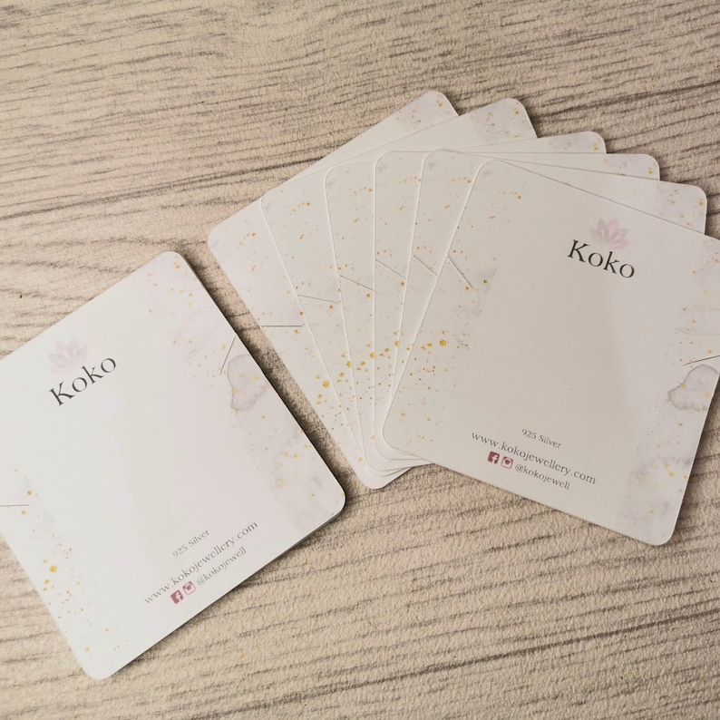K&ouml;nnte beinhalten: Ein Stapel wei&szlig;er Visitenkarten mit goldenen Flocken. Die Karten tragen den Schriftzug "Koko" in Schwarz. Der Text "925 Silver" und "www.kokojewellery.com" ist ebenfalls auf den Karten gedruckt, sowie Social-Media-Icons.