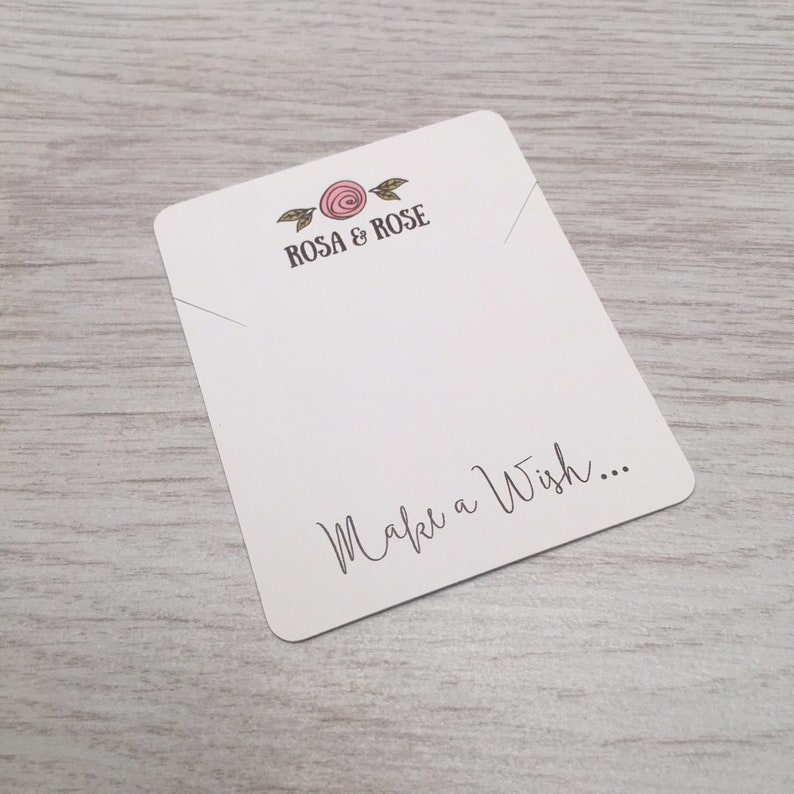 K&ouml;nnte beinhalten: Eine wei&szlig;e Karte mit einem Design aus einer rosa Rose und gr&uuml;nen Bl&auml;ttern. Der Text "ROSA & ROSE" ist in Schwarz gedruckt. Der Text "Make a Wish..." ist in schwarzer Kursivschrift gedruckt.