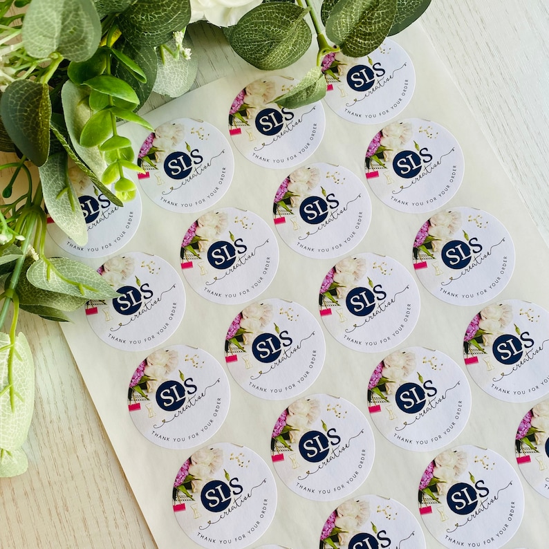 Op de afbeelding: Een vel met witte ronde stickers met een bloemenmotief en de tekst "SLS creative" en "Bedankt voor uw bestelling".