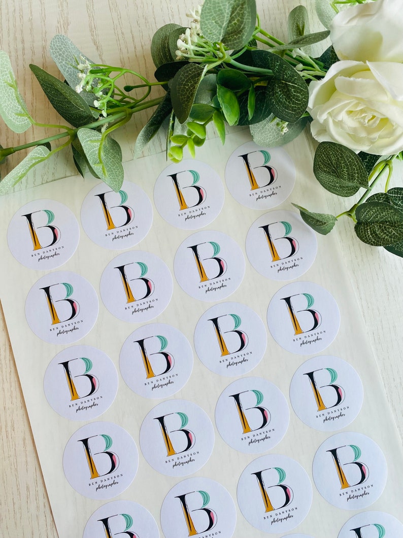 Op de afbeelding: Een vel met witte ronde stickers met een zwarte letter B, een geel en roze ontwerp en de tekst "Ben Dantzon Photographer" die op elke sticker wordt herhaald.
