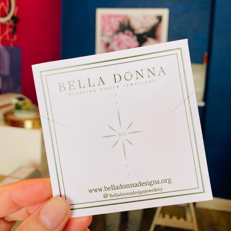 K&ouml;nnte beinhalten: Eine wei&szlig;e Karte mit goldener Beschriftung, auf der "Bella Donna Sterling Silver Jewellery" steht. Die Karte zeigt au&szlig;erdem einen stilisierten Stern mit den Buchstaben "BD" in der Mitte. Die Website-Adresse "www.belladonnadesigns.org" und der Social-Media-Handle "@belladonnadesignjewellery" sind unten auf der Karte aufgedruckt.