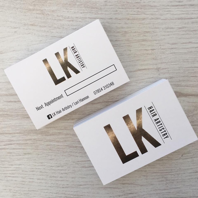 May include: Two white business cards with gold foil lettering. The text on the cards reads "LK Hair Artistry" and includes a phone number. The cards are stacked on top of each other.
