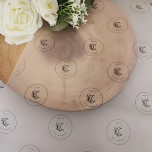 May include: A wooden round serving board with a light brown finish, overlaid with semi-transparent paper printed with a circular logo featuring a crescent moon and floral design. White roses and greenery are arranged on the board.