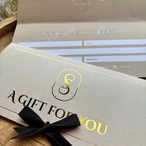 Op de afbeelding: Een cadeaubon met een zwarte strik. De bon is lichtgrijs met gouden letters met de tekst "A GIFT FOR YOU". Het Skin Sculpt Studio-logo is ook in goudfolie. De achtergrond is lichtbruin.