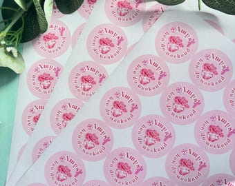 May include: Sheets of pink circular stickers with the text "Amys hen do weekend" in a white script font. Each sticker features a pink floral design. The stickers are arranged on a white surface.