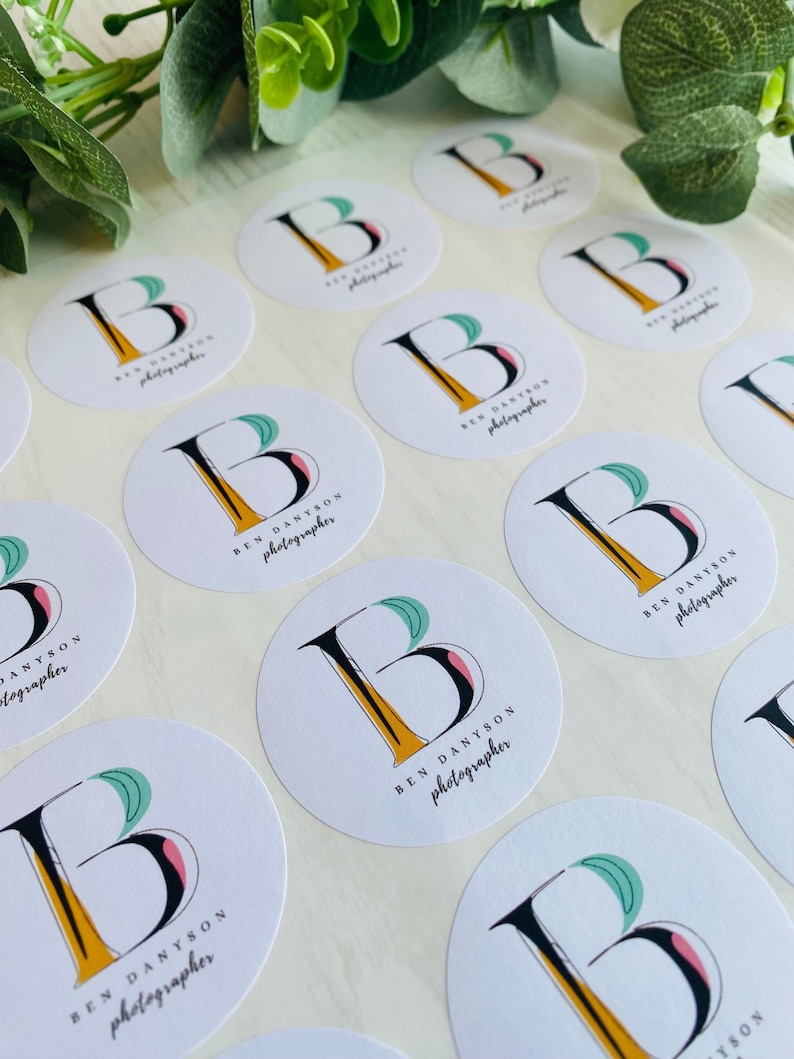 Op de afbeelding: Een vel met ronde witte stickers met een zwart en geel letter B logo en de tekst "BEN DANYSON fotograaf" gedrukt op elke sticker.