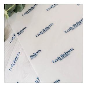 May include: White tissue paper sheets with a repeating pattern of the text "Leah Roberts Photography" in navy blue. The paper is slightly translucent, with a few sheets overlapping. A sprig of greenery is visible in the upper left corner.