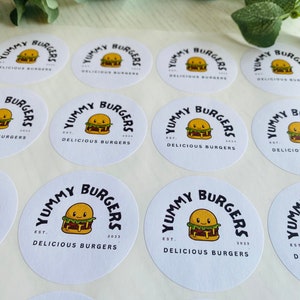 Op de afbeelding: Een vel met witte ronde stickers met een cartoon burgerontwerp. De stickers zeggen "Yummy Burgers Est. 2023 Delicious Burgers".