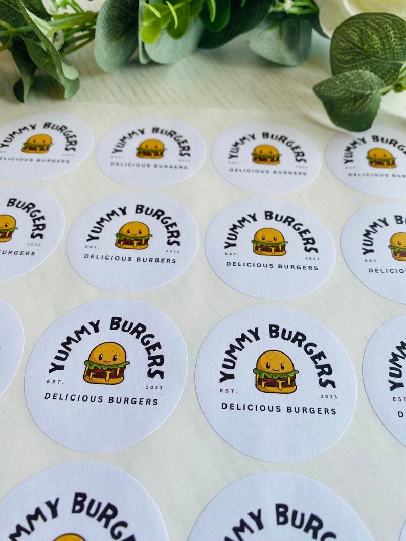 Op de afbeelding: Een vel met witte ronde stickers met een cartoon burgerontwerp. De stickers zeggen "Yummy Burgers Est. 2023 Delicious Burgers".