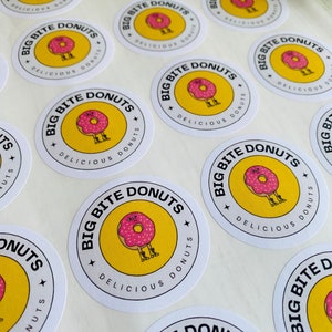 Op de afbeelding: Een verzameling ronde stickers met een gele achtergrond en zwarte tekst die "Big Bite Donuts" en "Delicious Donuts" luidt. Een cartoon-donut met roze glazuur en sprenkels draagt laarzen en heeft een gezicht.